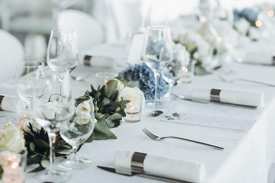 Special Event Insurance - Wedding Table Decoration with Floral Garland and Blue Flowers Between Glasses on a White Tablecloth