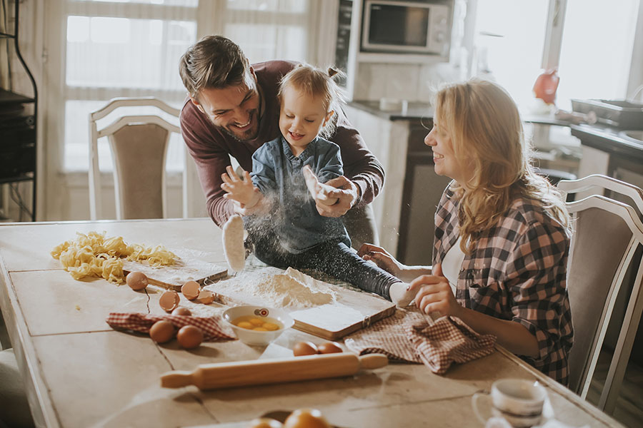 Personal Insurance - Happy Family Making Pasta in a Kitchen at Home With Young Child Sitting on the Kitchen Table and Parents Laughing